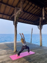 Una persona realizando una postura de yoga sobre una esterilla rosa debajo de una estructura de madera, con vista al océano.