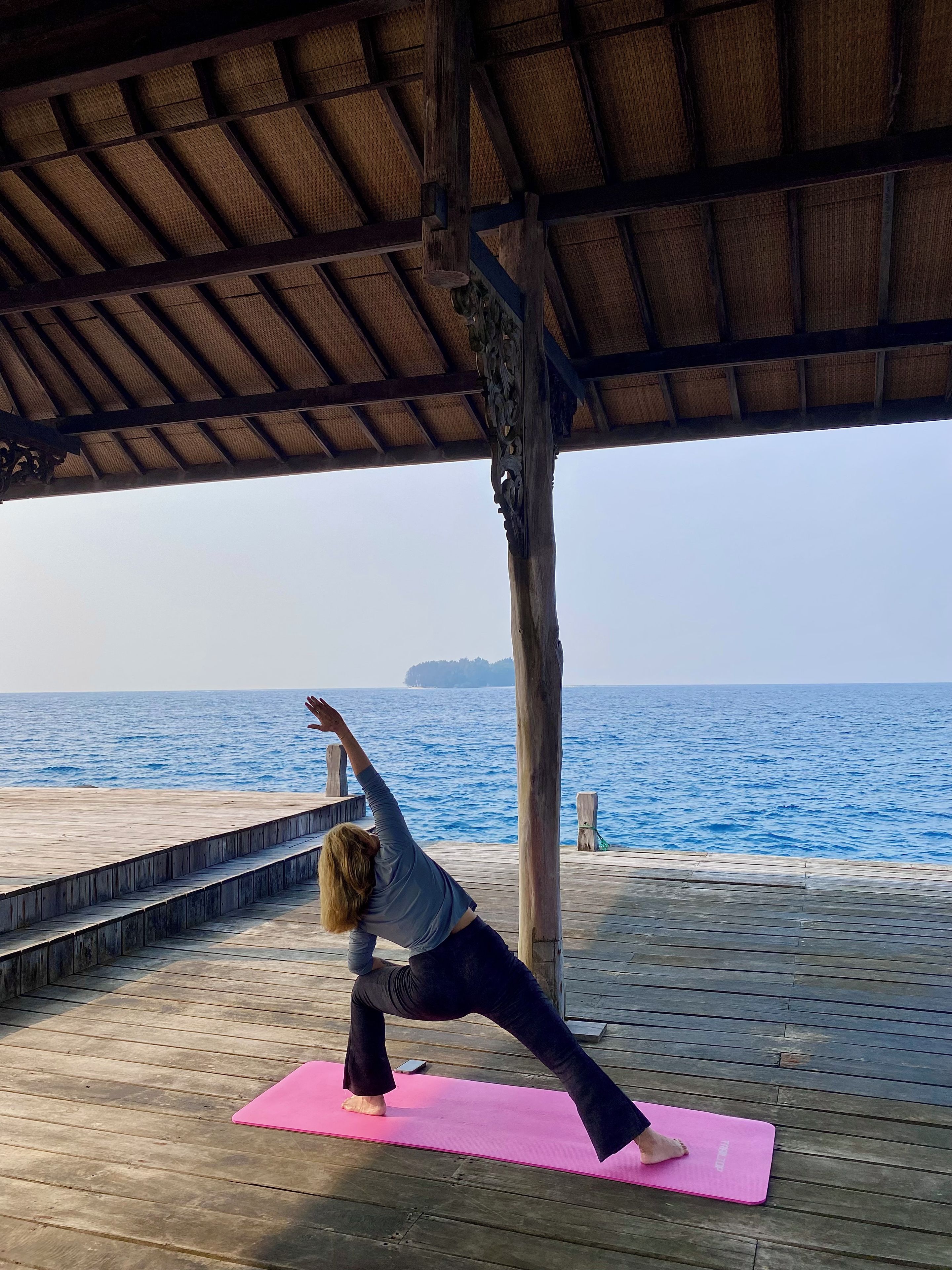 Eine Person führt eine Yogaübung auf einer rosafarbenen Matte unter einer Holzbeschaffenheit mit Blick auf das Meer aus.