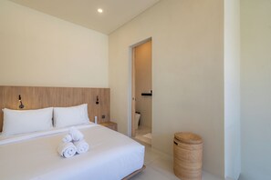 A modern hotel room with a large bed, a wooden headboard, a bedside table, a waste basket, and a door leading to a bathroom. - Villa Naya by Betterplace (Pecatu)