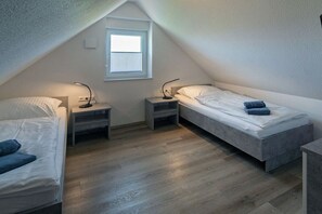 A small, triangular attic bedroom with two beds, a window, and a nightstand with a lamp. - Holiday home with 1 bedroom (Bensersiel)