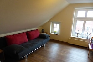 Ein Zimmer mit einem dunkelgrauen Sofa, einem kleinen Tisch mit einer Lampe, einem Fenster mit Blick auf Gebäude und einem Heizkörper unter dem Fenster.