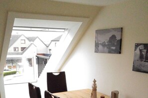 Un espace repas comprenant une table, des chaises et une vue sur les maisons voisines, visible par une fenêtre.