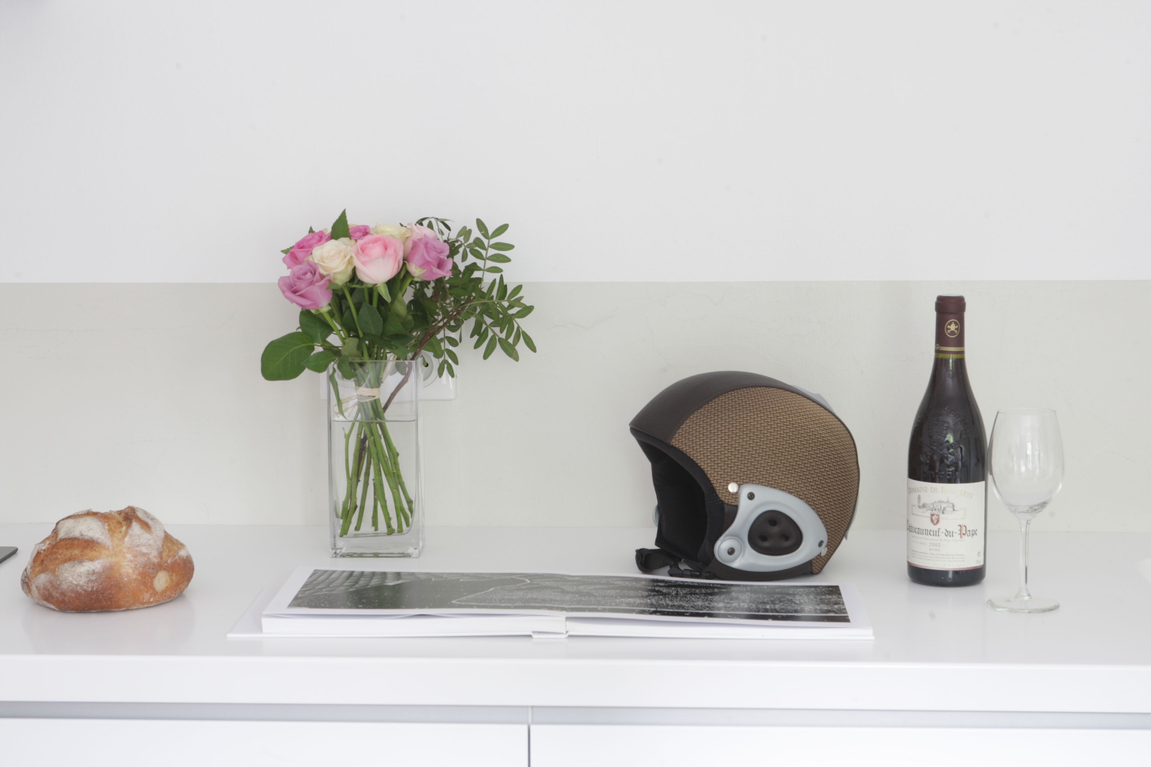 A white counter with a vase of pink flowers, a helmet, a bottle of wine, and a glass.