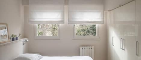 Une chambre à coucher avec un lit, deux fenêtres équipées de stores, un radiateur et une armoire.
