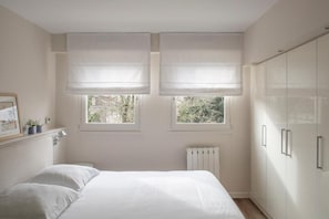 A bedroom with a bed, two windows with blinds, a radiator, and a wardrobe. - Les Suites de Catherine, la si Calme (Nancy)