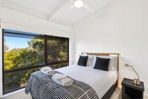A bedroom with a large window, a bed with a patterned bedspread, two pillows, a bedside table with a lamp, and a ceiling fan. - Ocean View House Fairhaven (Fairhaven)