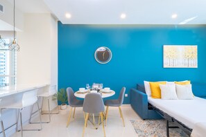 Ein modernes Zimmer mit blauer Akzentwand, einer runden Uhr, einem Esstisch mit Stühlen, einer Couch mit Kissen und einem Bett.