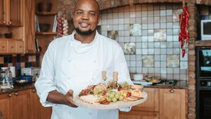 A chef holding a platter of food in a kitchen with wooden cabinets and a tiled backsplash. - Sandcastle (Wesley)