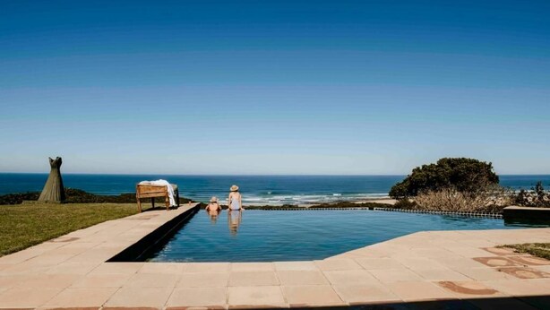 A swimming pool with a view of the ocean, a bench, and a small tree. - Sandcastle (Wesley)