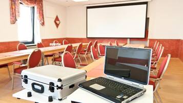 A conference room with a projector screen, tables, chairs, a laptop, and a suitcase.