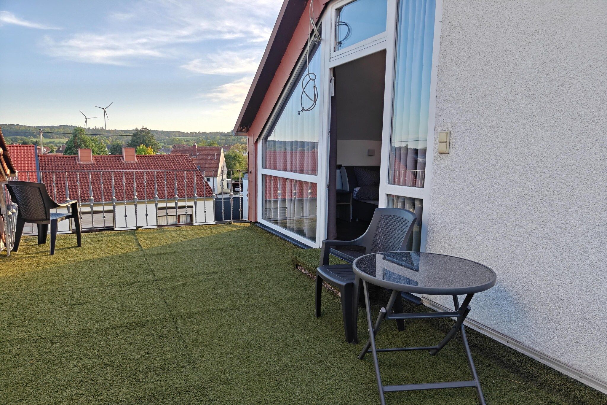 Ein Wohnhaus mit einer Patio, ausgestattet mit zwei Stühlen und einem kleinen Tisch, einer Glastür und Blick auf ein Gebäude mit rotem Ziegeldach im Hintergrund.