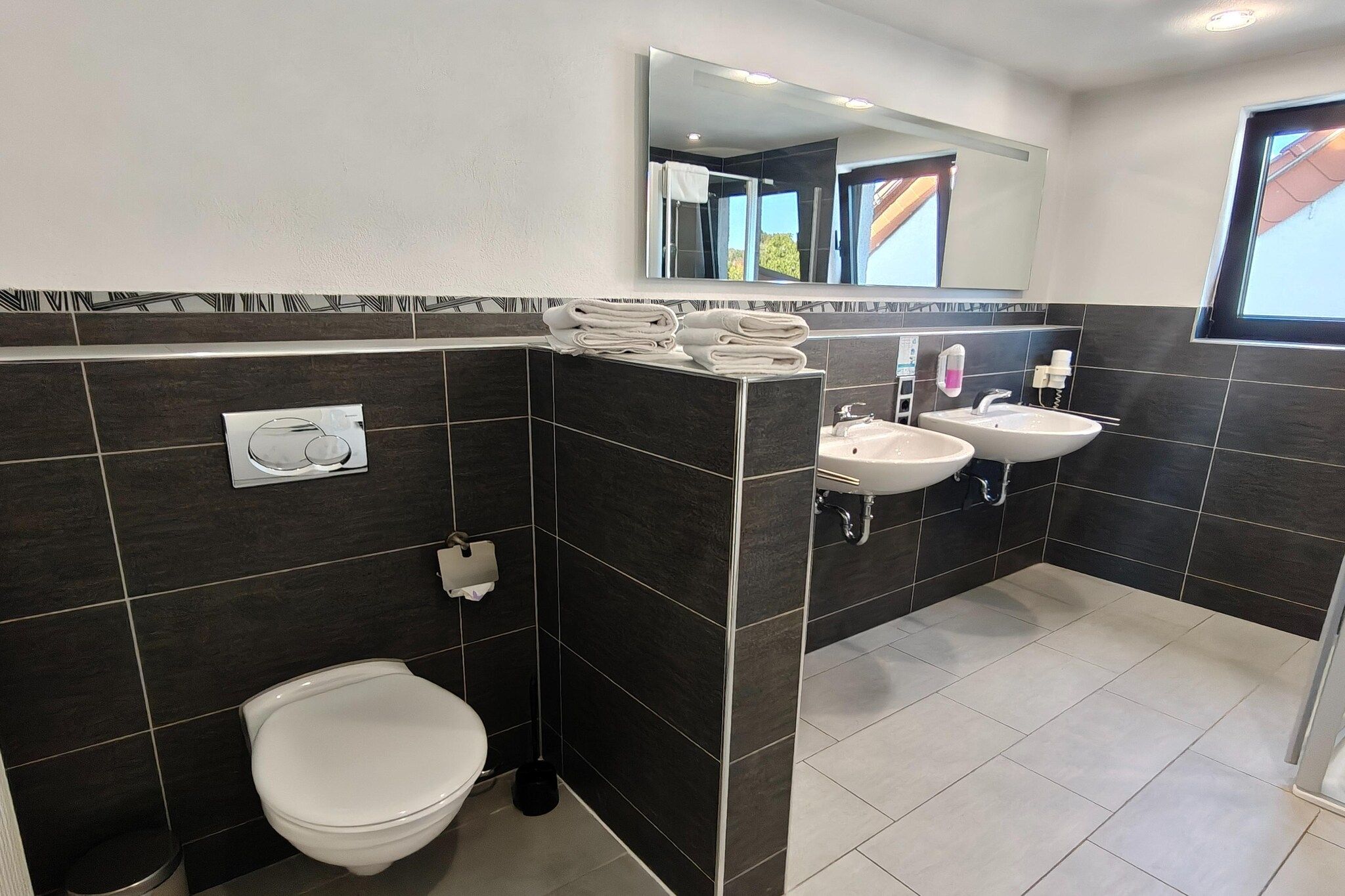 Un baño con inodoro, dos lavamanos, toallero con toallas, espejo y ventana.