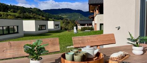 Un espacio de comedor al aire libre con una mesa de madera, plantas en macetas y vistas a edificios modernos y montañas.