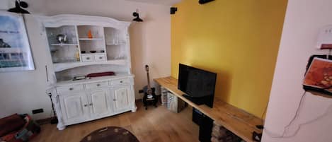 Un salon avec un téléviseur posé sur un meuble en bois, une vitrine blanche contenant de la verrerie, une guitare et un fauteuil rouge.