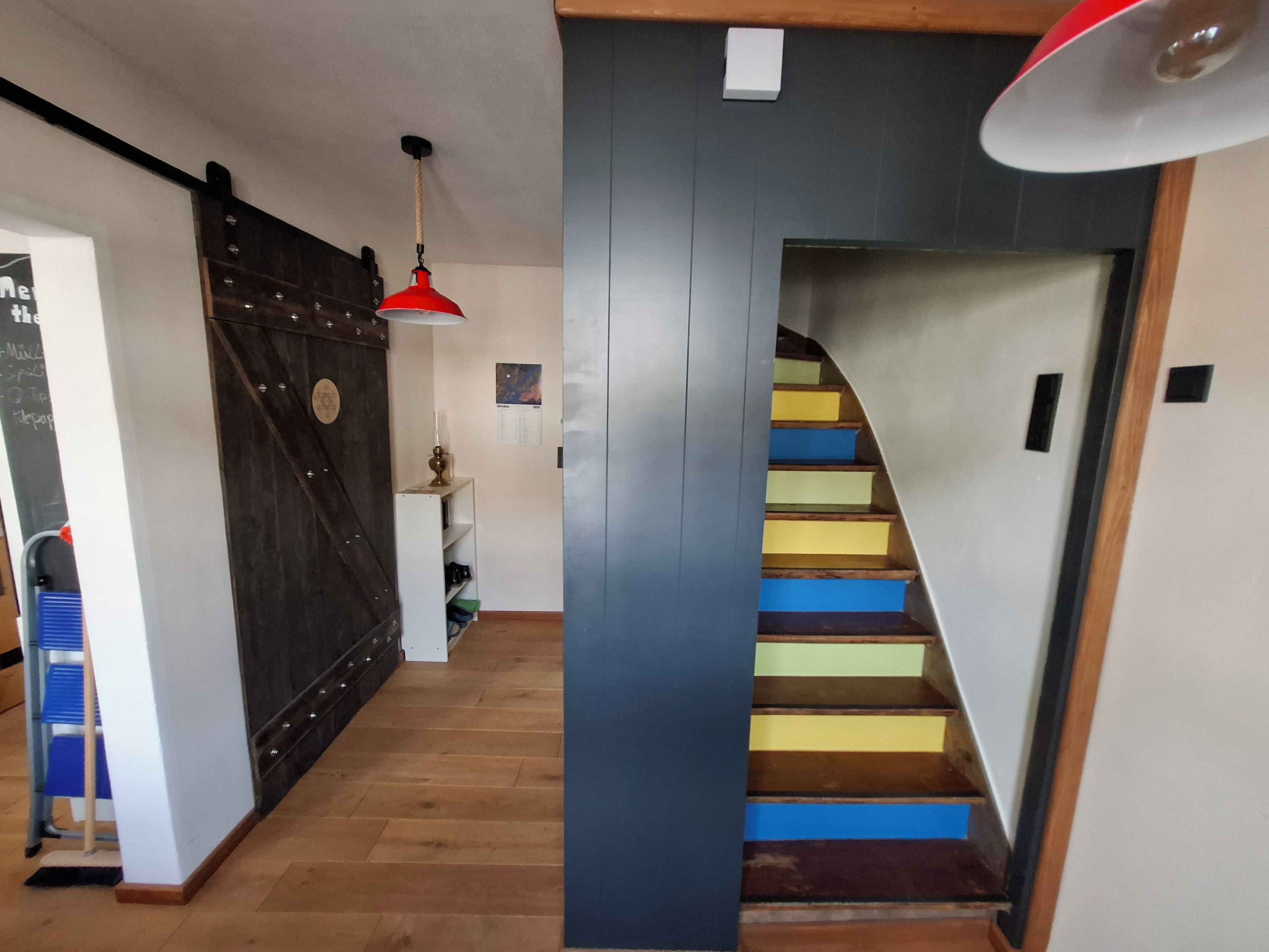 A staircase with colorful steps leading to a landing, a dark wooden door, and a white wall with a small shelf and a lamp.