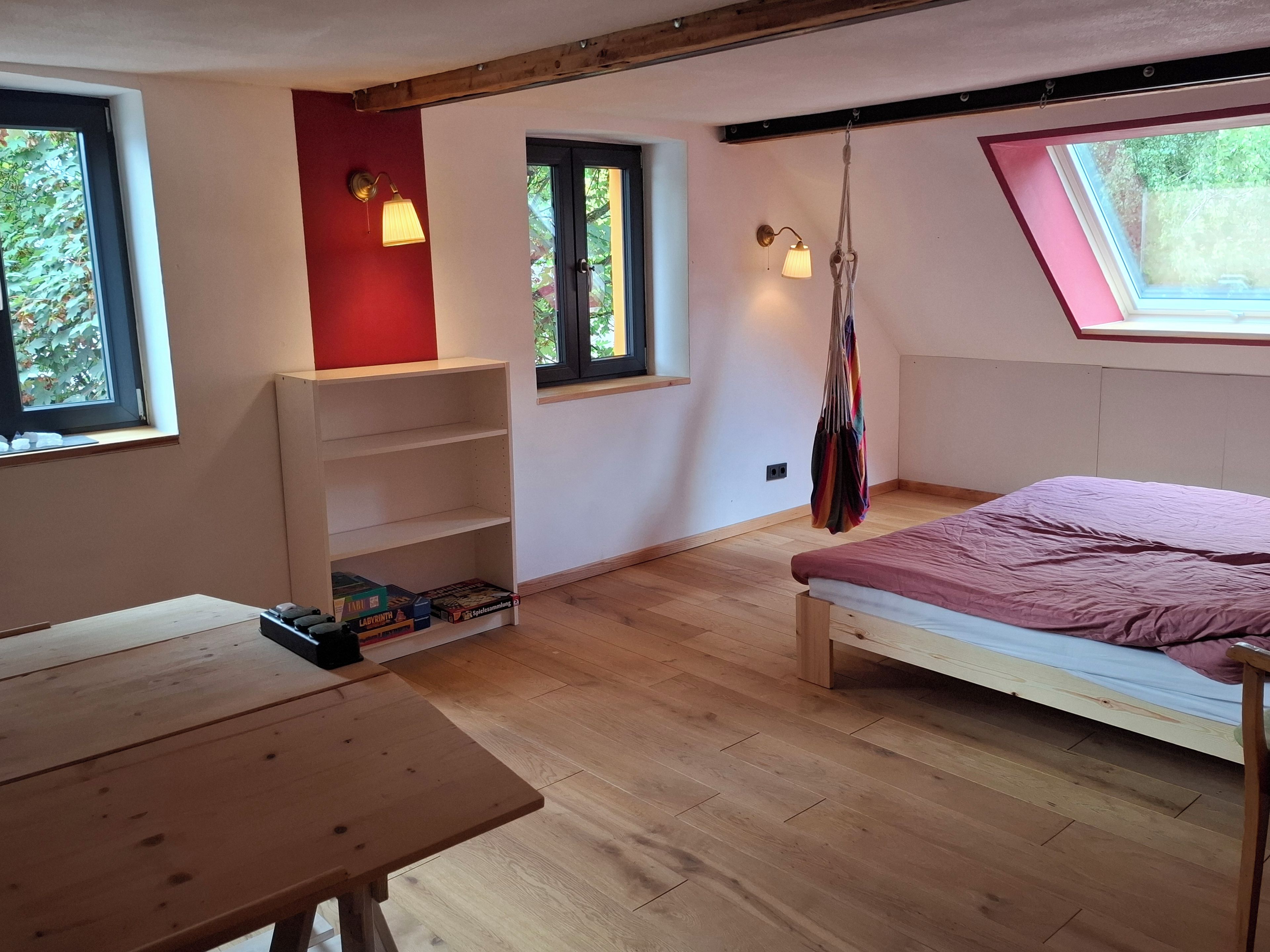 Ein Zimmer mit einem Bett, einem Tisch und einer Regaleinrichtung. Es gibt Fenster und ein Dachfenster.