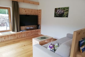 A living room with a flat-screen TV mounted on a wooden wall, a sofa, a coffee table, and a stone wall visible through a window. - At Niedermoar- "Etschwool" (Naturns)