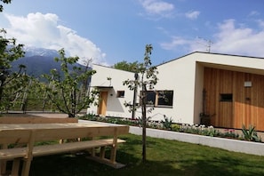 A modern building with a wooden door, a bench, and a garden with trees and flowers. - At Niedermoar- "Etschwool" (Naturns)