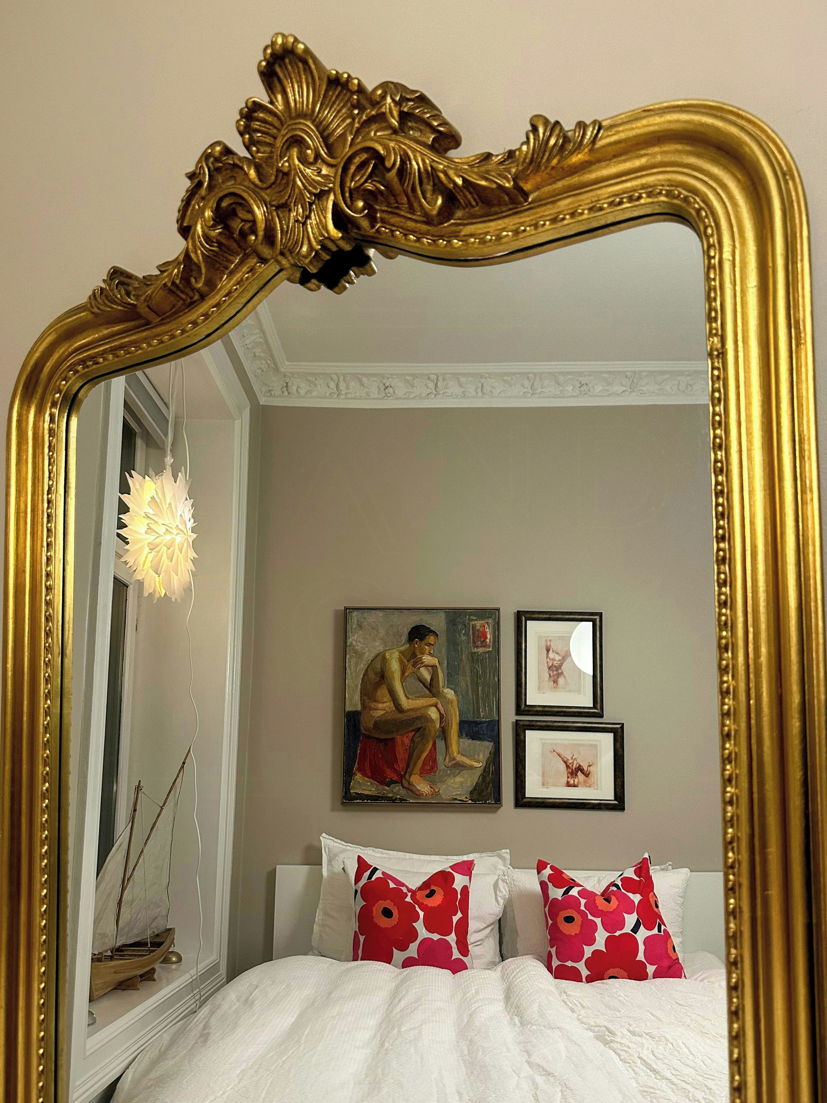 Un dormitorio con un espejo grande y ornamentado, una cama con ropa de cama blanca y dos cuadros en la pared.