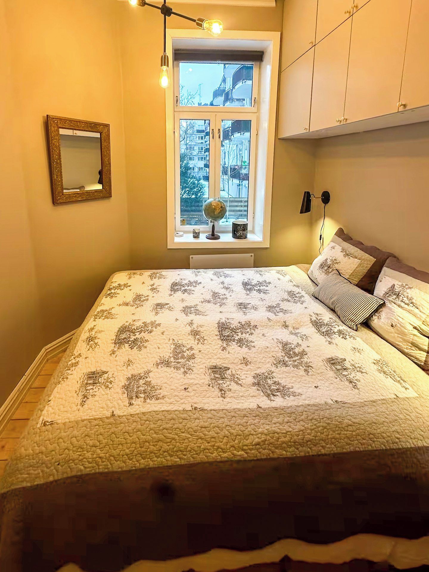 Een slaapkamer met een bed, een raam met uitzicht op gebouwen, een wereldbol en een lamp.