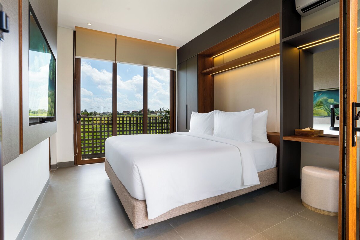 A modern hotel room with a large bed, a television mounted on the wall, and a view of a green landscape through the window.