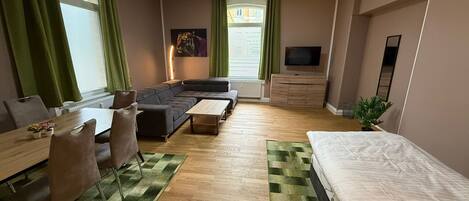 Una habitación de hotel moderna con sofá, una mesa de comedor con sillas, cama, televisor y ventana con cortinas.