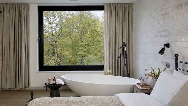Een slaapkamer met een groot raam, een badkuip, een bed en een nachtkastje met een lamp.