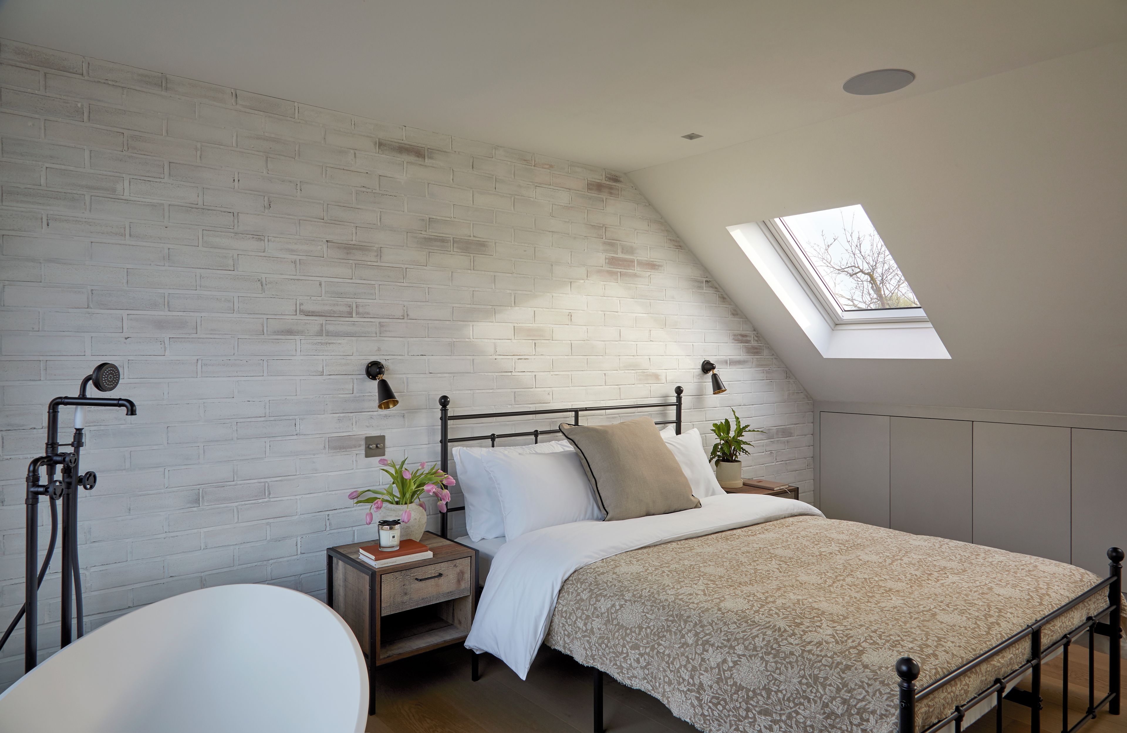 Una camera da letto con soffitto spiovente, un lucernario, un letto con copriletto a motivi, un comodino con una pianta e una sedia bianca.