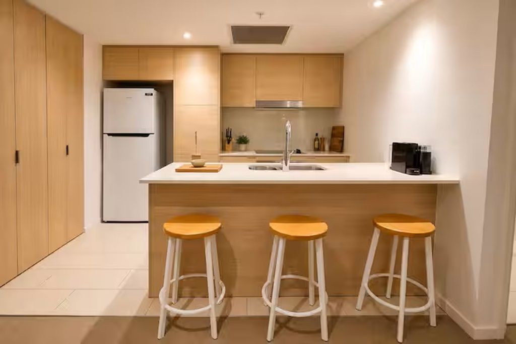 A modern kitchen with a central island, three stools, a refrigerator, and a coffee machine.