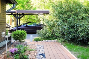 Eine Holzterrasse führt zu einem überdachten Parkplatz, umgeben von üppiger Vegetation und einem kleinen Garten mit violetten Blüten.