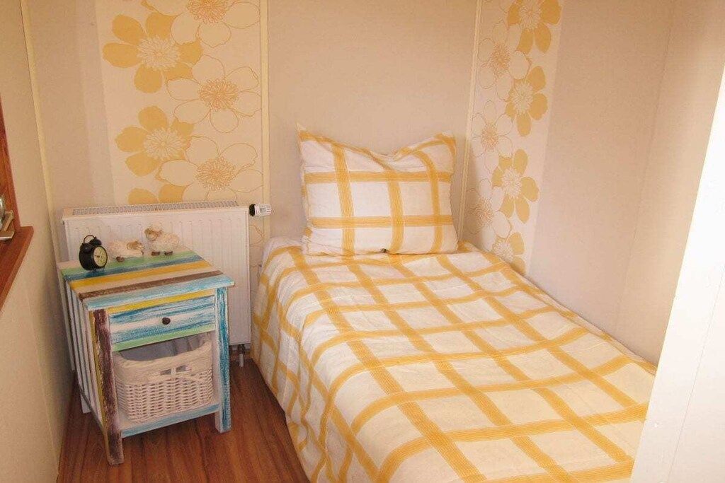 Ein kleines Zimmer mit einem Einzelbett, einer Bettdecke mit Schachmuster, einem Nachttisch mit einer Uhr und einer Wand mit Blumenpapier.