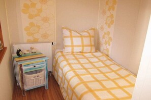 A small, single-bed room with a checkered bedspread, a bedside table with a clock, and a wall with floral wallpaper. - Heideblockhaus Dachsbau (Neuenkirchen)