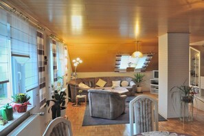 Ein modernes Wohnzimmer mit einer Couch, einem Couchtisch und einem Essbereich mit Stühlen. Es gibt Pflanzen und eine Lampe.