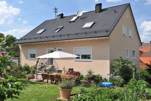 A two-story house with a sloped roof, a patio with a table and chairs, and a garden with various plants and flowers. - Holiday apartment with 2 bedrooms (Egloffstein)