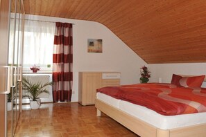 A bedroom with a wooden ceiling, a bed with a red and white bedspread, a wooden nightstand, a white radiator, and a window with curtains. - Holiday apartment with 2 bedrooms (Egloffstein)