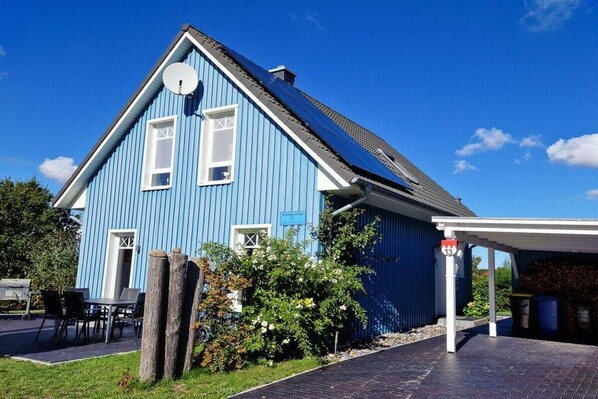 A blue building with a satellite dish, a covered parking area, and a small garden with flowers. - Holiday home in Bodstedt with swimming pool (Bodstedt)