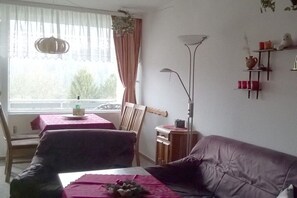 Ein Wohnzimmer mit einer Couch, einem Esstisch mit Stühlen, einer Stehlampe und einem Fenster mit Vorhängen.