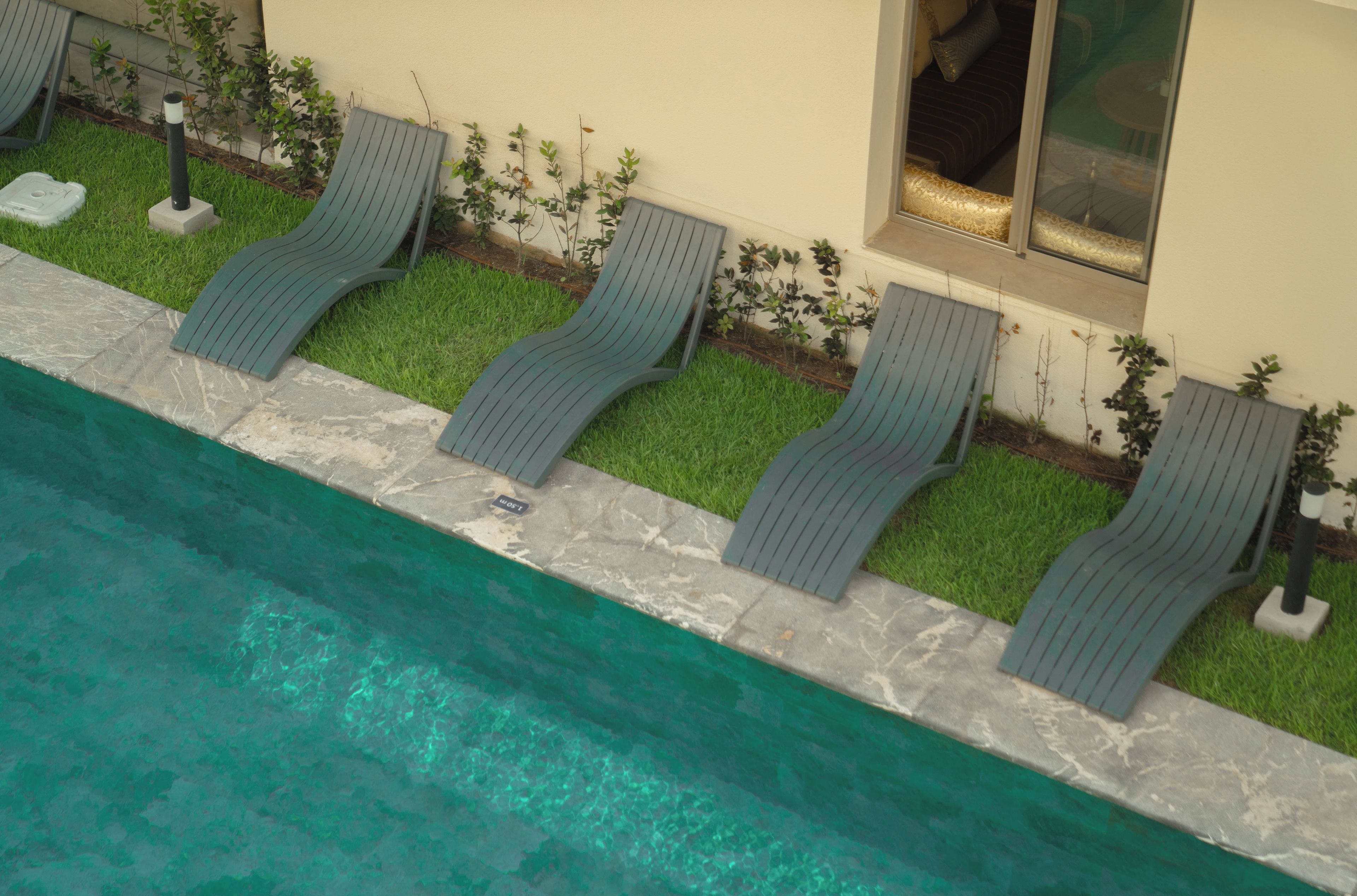 Un área junto a la piscina con sillones de descanso a lo largo del borde, un pequeño jardín con arbustos y un edificio con una ventana.