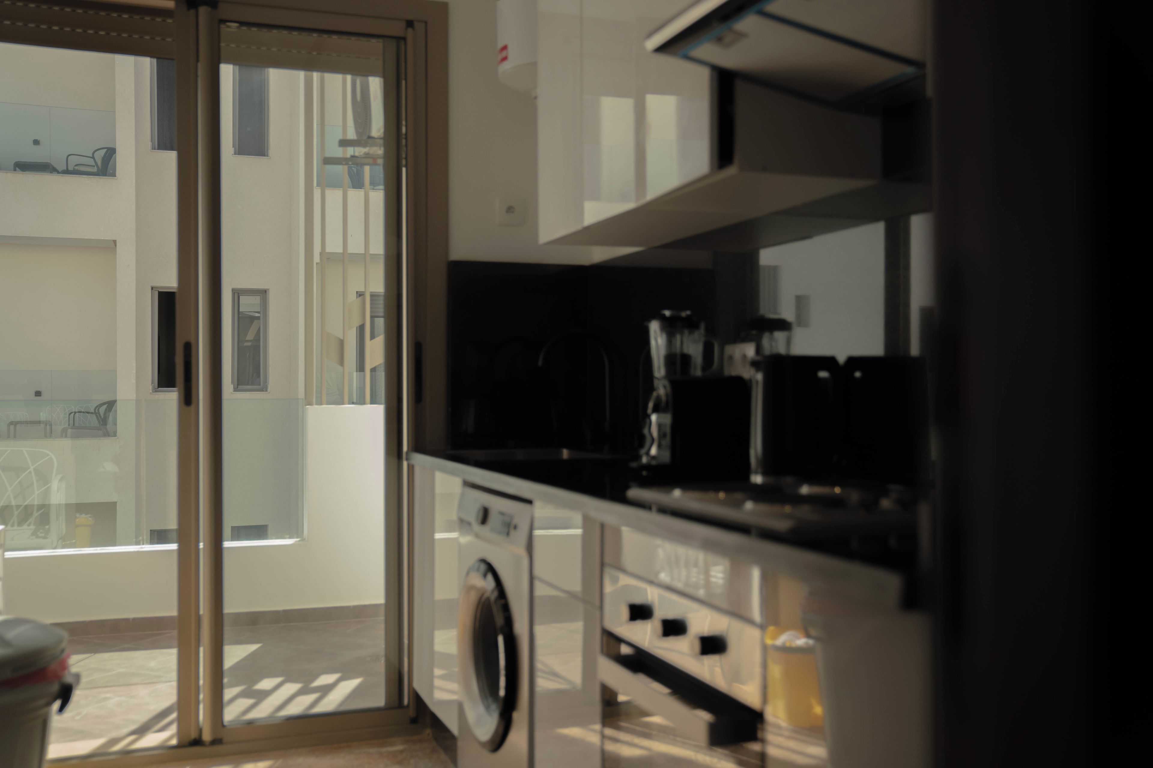 Cozinha moderna com máquina de lavar embutida, cafeteira e vista para uma varanda através de uma porta de vidro.