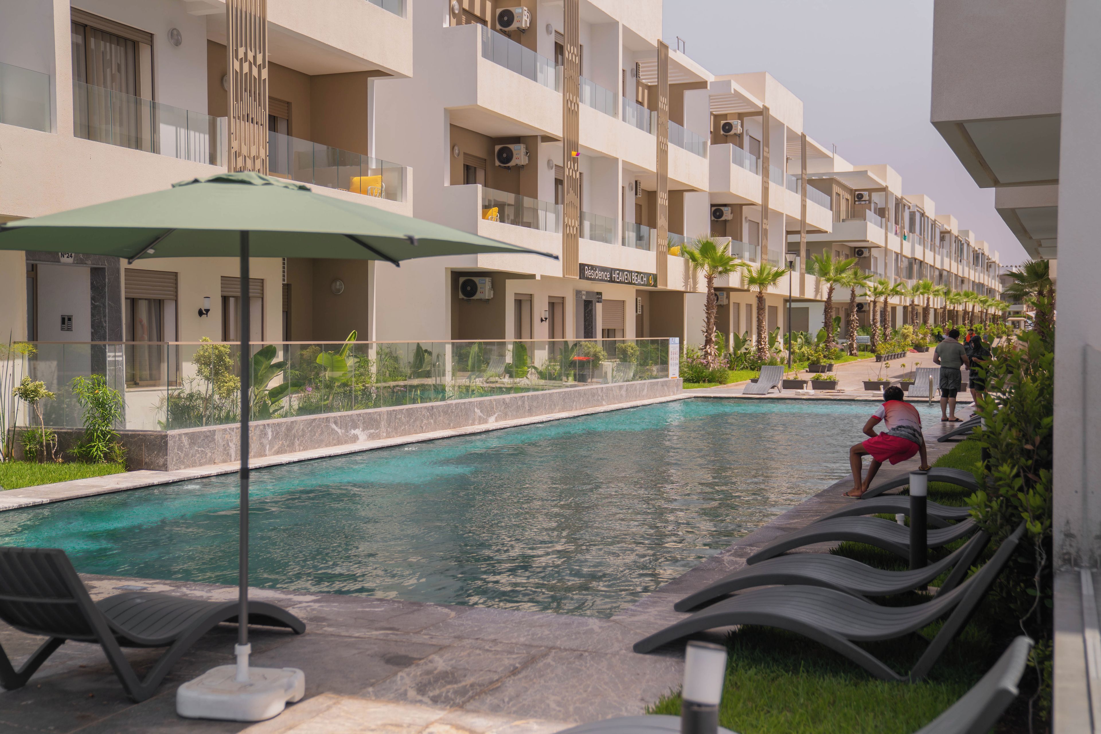 A swimming pool with lounge chairs and a palm tree, surrounded by a building with balconies and air conditioning units.