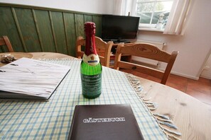 Eine Flasche Champagner, eine karierte Tischdecke und ein Buch mit dem Titel 'Gästebuch' auf einem Holztisch in einem Raum mit Fernseher und Fenster.