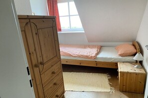 A small, single-bed room with a sloped ceiling, a wooden wardrobe, a bedside table with a lamp, and a window with curtains. - Holiday apartment with 3 bedrooms (Tönning)
