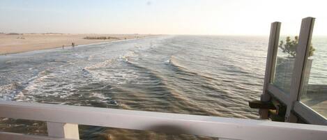 A beach view from a wooden deck with a railing, ocean waves, and a distant shoreline.