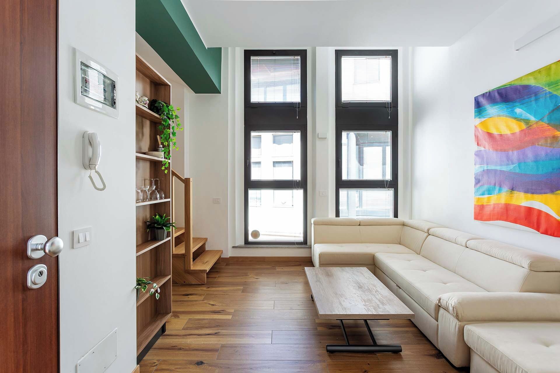 Un soggiorno moderno con un divano color beige, un tavolino in legno e una libreria con piante. Sono presenti due ampie finestre con persiane e un quadro astratto dai colori vivaci appeso alla parete.