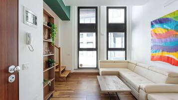 Un soggiorno moderno con un divano color beige, un tavolino in legno e una libreria con piante. Sono presenti due ampie finestre con persiane e un quadro astratto dai colori vivaci appeso alla parete.