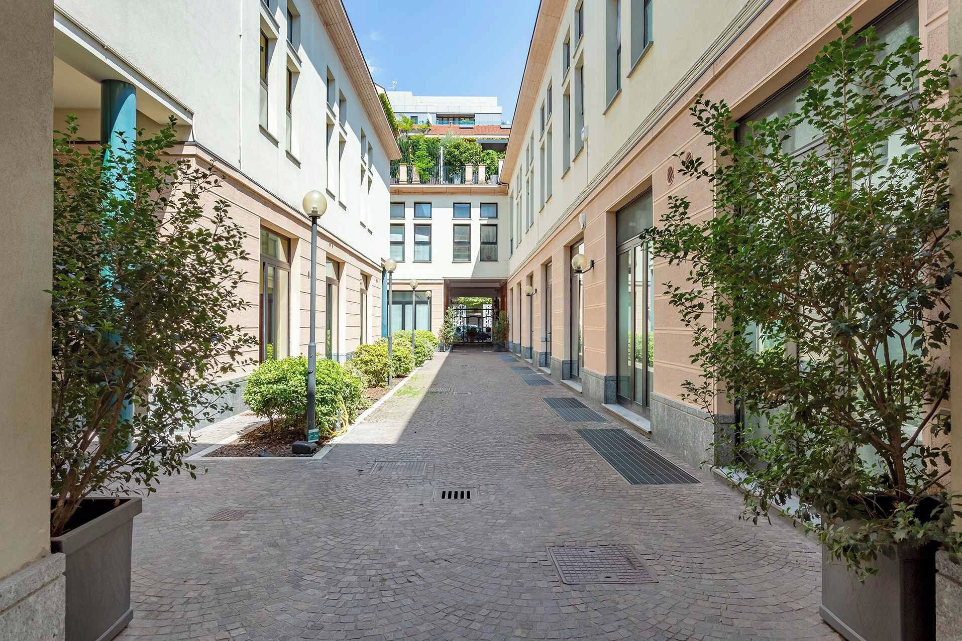 A narrow, paved corridor with buildings on either side, lined with small trees and street lamps.