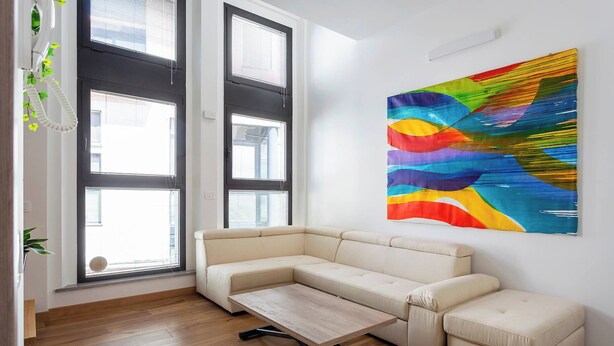 Ein modernes Wohnzimmer mit einem beigefarbenen Ecksofa, einem Couchtisch aus Holz und einem farbenfrohen, abstrakten Gemälde an der Wand.