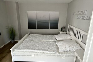 A bedroom with a white bed, a nightstand, a wall-mounted artwork, and a potted plant. - Holiday apartment with 1 bedroom (Cuxhaven)