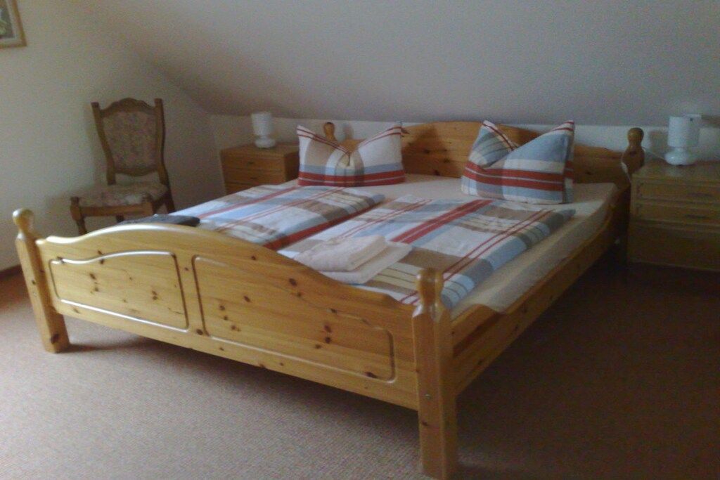 Un lit en bois avec une couverture à carreaux et des oreillers, une table de chevet en bois avec une lampe, et une chaise dans une pièce dont le sol est recouvert d’une moquette beige.