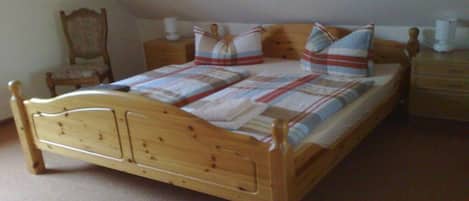 A wooden bed with a plaid bedspread and pillows, a wooden nightstand with a lamp, and a chair in a room with beige carpet.
